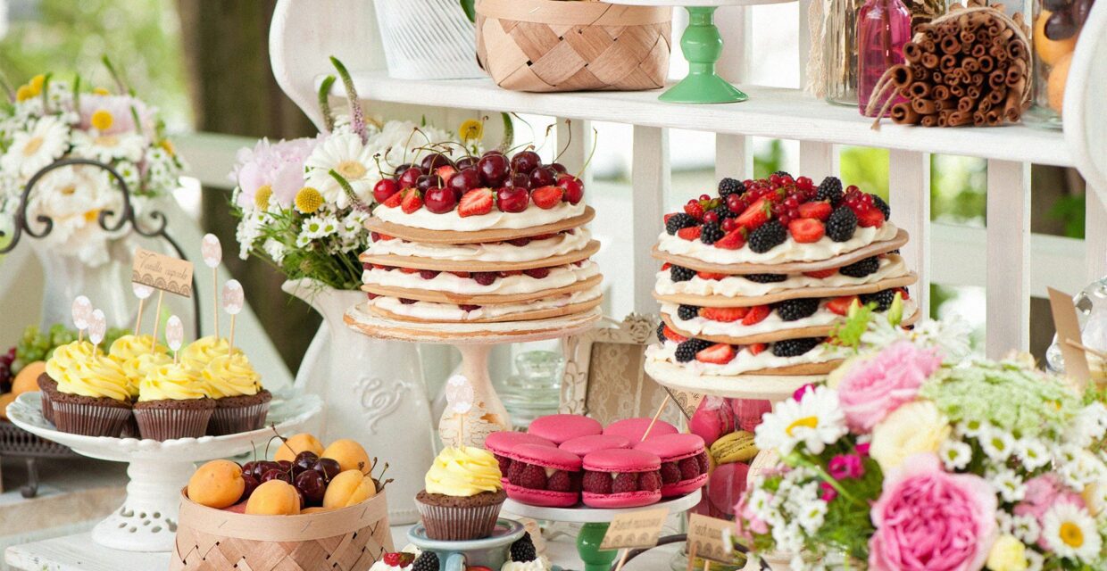 Elegant dessert table with layered fruit cakes, cupcakes, pink macarons, fresh berries, flowers, and assorted sweets for a catered celebration.