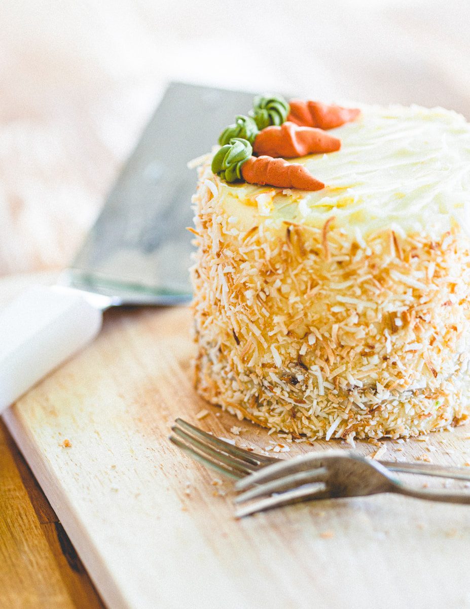 Mini carrot cake covered in toasted coconut flakes and topped with cream cheese frosting and decorative buttercream carrots on a wooden board.