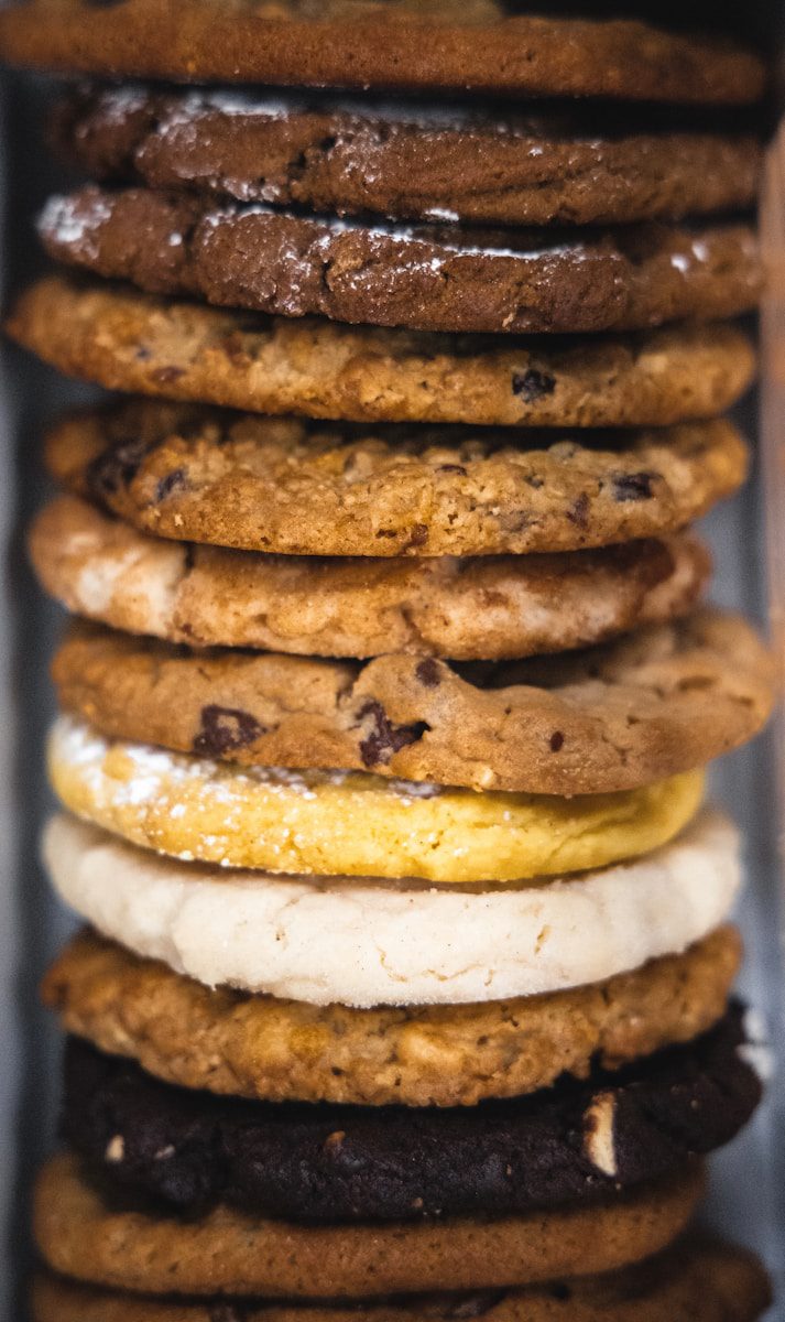 Assorted stack of freshly baked gourmet cookies featuring chocolate chip, double chocolate, sugar, lemon, oatmeal, and specialty cookies displayed in a bakery-style assortment.