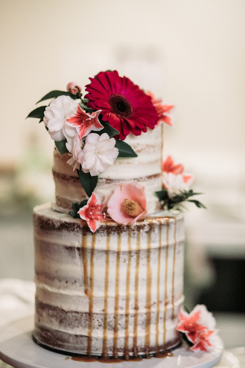 Rustic semi-naked two-tier wedding cake with caramel drip, decorated with fresh pink, white, and coral flowers, including a vibrant gerbera daisy, displayed on a white cake stand.