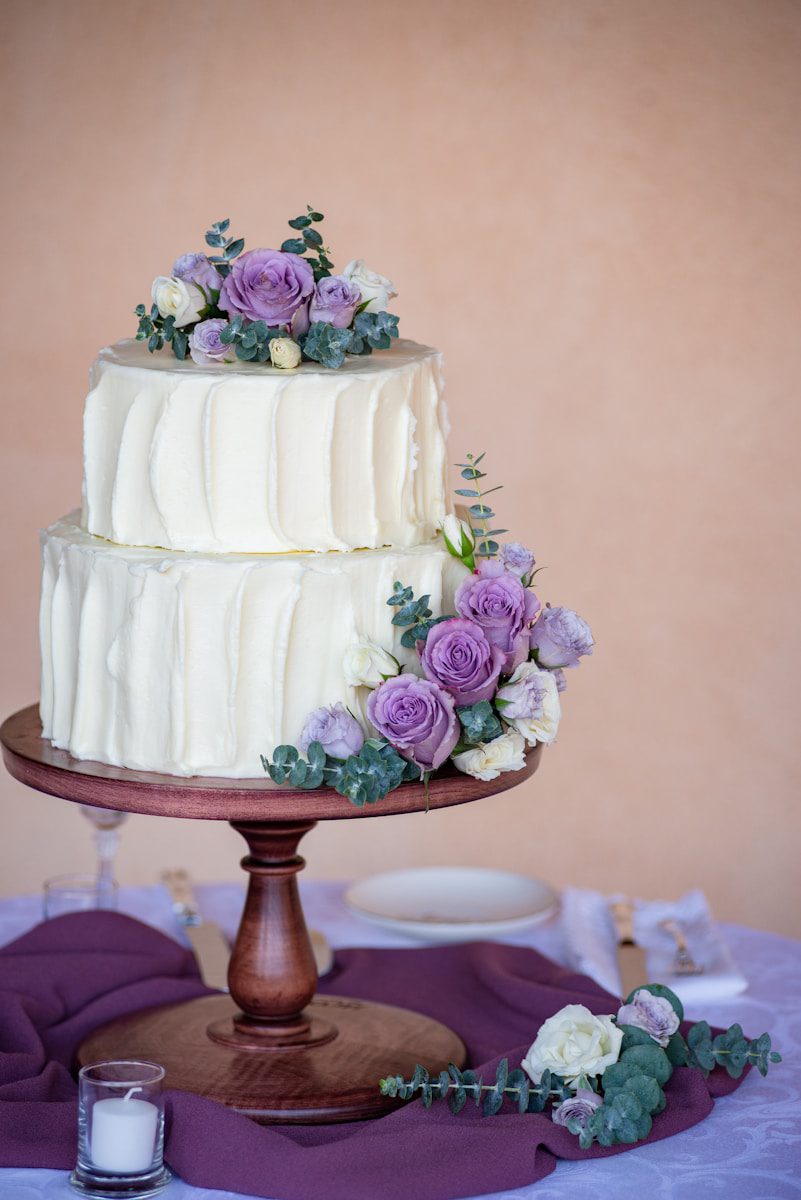Elegant two-tier white wedding cake with textured buttercream frosting, decorated with lavender roses, white blooms, eucalyptus greenery, and displayed on a wooden cake stand.