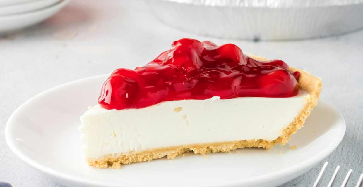 Classic New York-style cheesecake slice with a buttery graham cracker crust topped with glossy cherry pie filling, served on a white dessert plate.