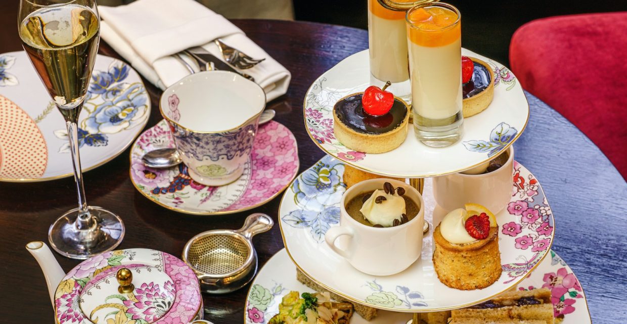 Elegant afternoon tea service featuring a three-tier stand with miniature desserts, finger sandwiches, smoked salmon canapés, tea, and sparkling wine served on floral china.