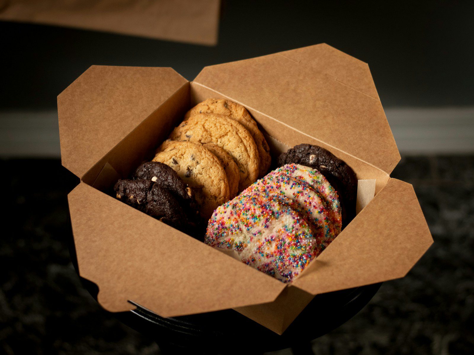 Assorted gourmet cookies including chocolate chip, double chocolate, and rainbow sprinkle sugar cookies neatly arranged inside a kraft bakery takeout box.