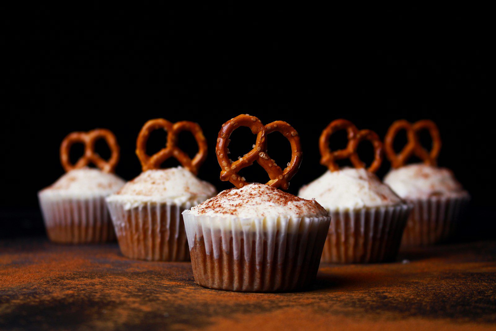 Vanilla cupcakes topped with cream cheese frosting, dusted with cinnamon, and garnished with mini pretzels, arranged against a dramatic black background.