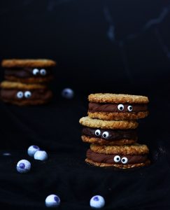 Halloween-themed chocolate cookie sandwiches filled with rich chocolate buttercream and decorated with candy eyes, stacked on a dark background with candy eyeball decorations.