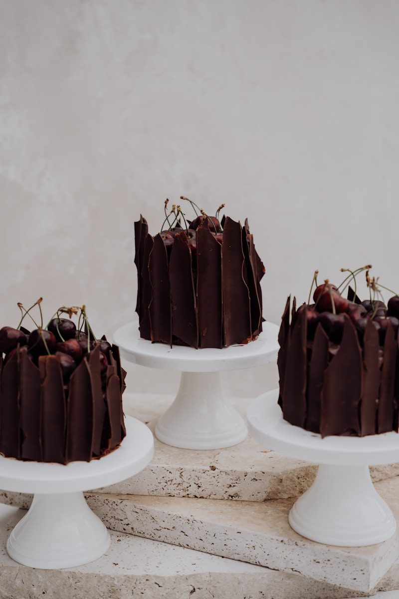 Individual dark chocolate cakes wrapped with handcrafted chocolate shards, topped with fresh cherries, and displayed on elegant white cake stands.
