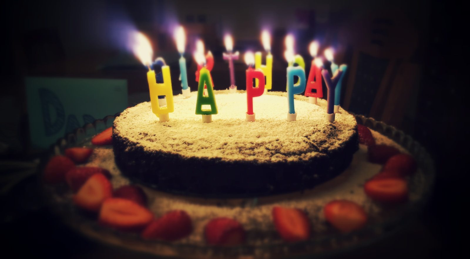 Chocolate birthday cake dusted with powdered sugar, topped with colorful "Happy Birthday" letter candles, and surrounded by fresh sliced strawberries.