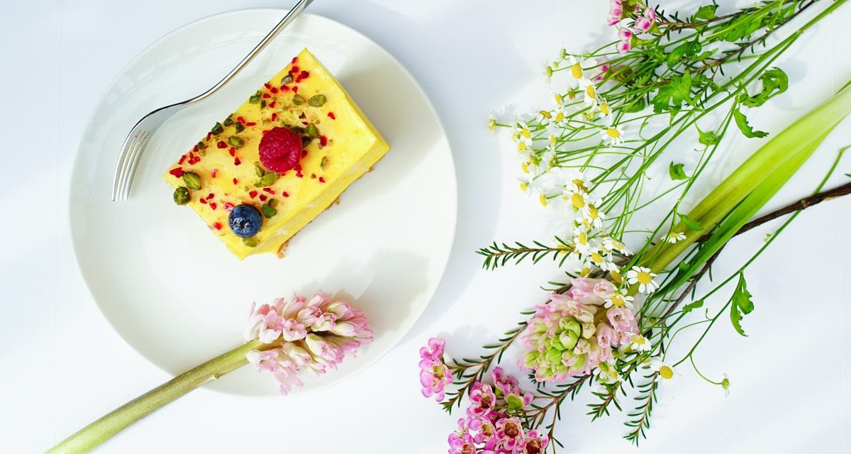 Elegant slice of layered vanilla cake topped with fresh raspberries, blueberries, pistachios, and edible flower petals, plated alongside a bouquet of fresh spring flowers.
