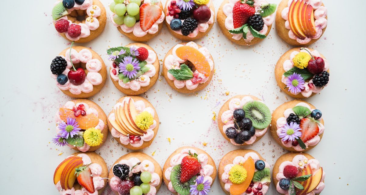 Assorted gourmet fruit tartlets topped with fresh strawberries, blueberries, blackberries, grapes, kiwi, peaches, edible flowers, and piped berry cream, beautifully arranged for an elegant dessert display.