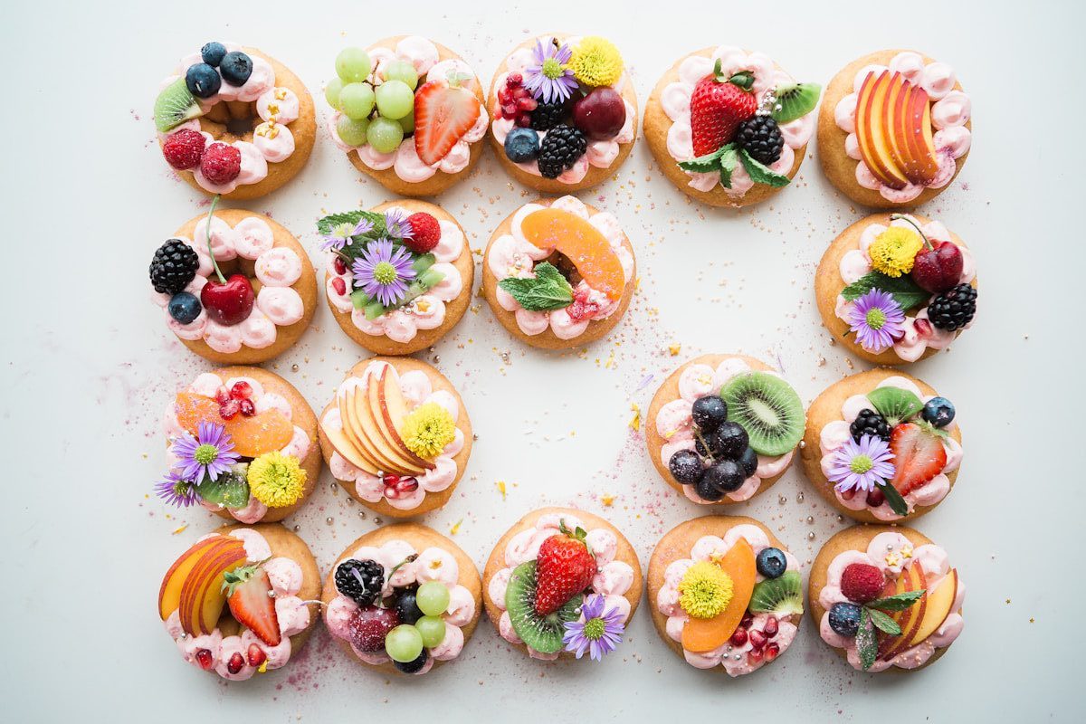 Assorted gourmet fruit tartlets topped with fresh strawberries, blueberries, blackberries, grapes, kiwi, peaches, edible flowers, and piped berry cream, beautifully arranged for an elegant dessert display.