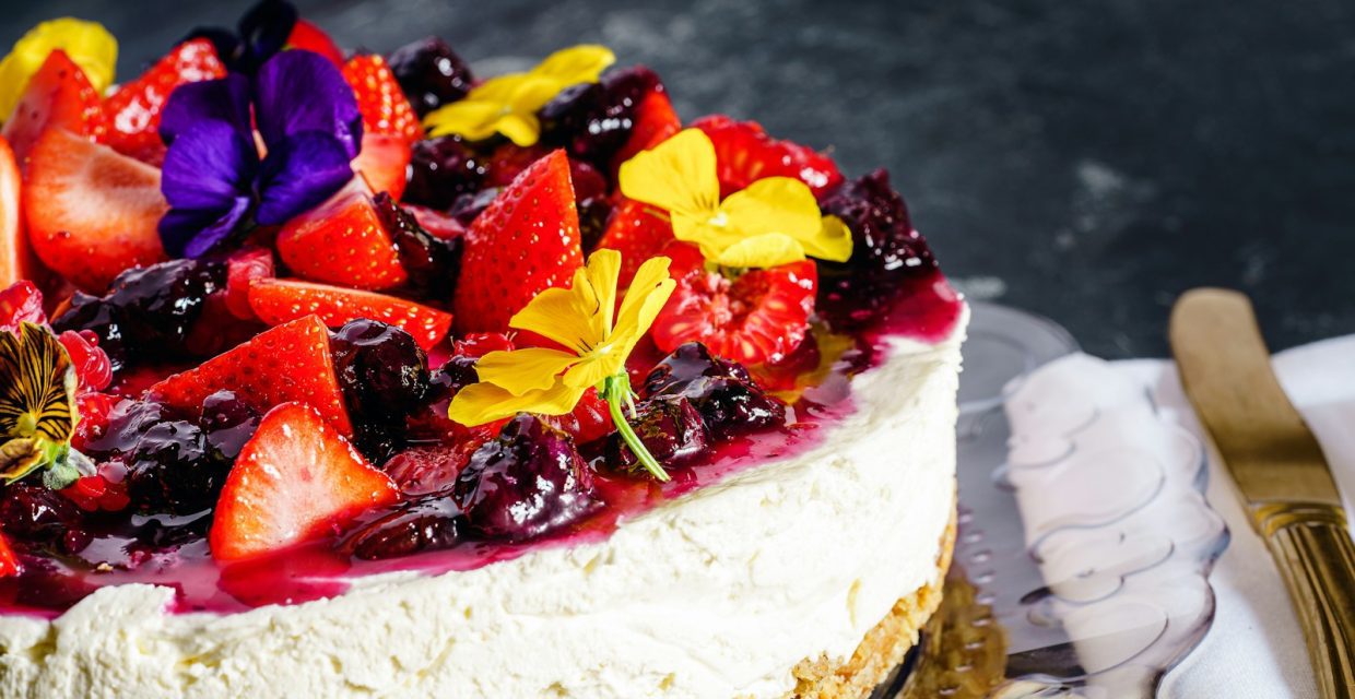 Creamy mixed berry cheesecake topped with fresh strawberries, blueberries, berry glaze, and colorful edible flowers on a graham cracker crust, perfect for elegant catered events in Charleston, SC.