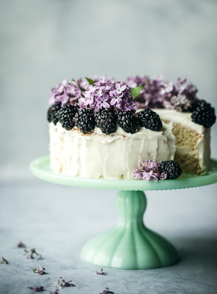 Rustic vanilla layer cake with smooth buttercream frosting, topped with fresh blackberries and edible lilac blossoms, displayed on a vintage cake stand for elegant catered events in Charleston, SC.