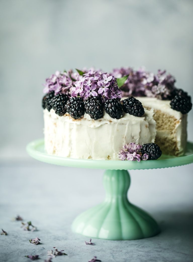 Rustic vanilla layer cake with smooth buttercream frosting, topped with fresh blackberries and edible lilac blossoms, displayed on a vintage cake stand for elegant catered events in Charleston, SC.