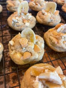 Soft homemade banana pudding cookies topped with vanilla wafer pieces, banana chips, white chocolate chips, and banana crumble, cooling on a wire rack.