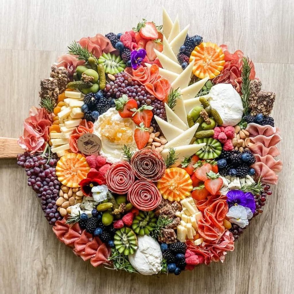 Luxury charcuterie board featuring artisan cheeses, handcrafted salami roses, prosciutto, burrata, fresh berries, grapes, kiwi, citrus, cornichons, nuts, edible flowers, and gourmet accompaniments by Charleston catering.