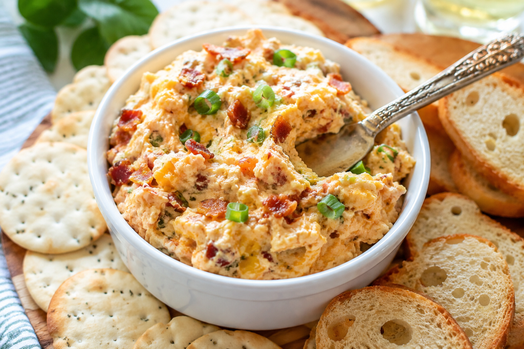 Creamy bacon cheddar cheese spread topped with green onions and crispy bacon, served with crackers and toasted bread.